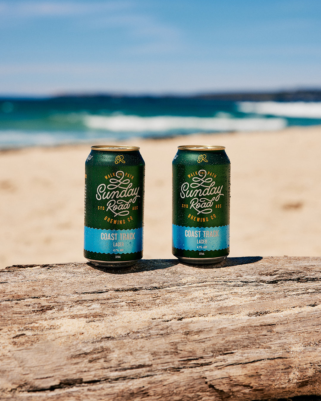 Two cans of Sunday Road Brewing coast track lager beer on a wooden log with a beach and ocean background in sydney