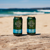 Two cans of Sunday Road Brewing coast track lager beer on a wooden log with a beach and ocean background in sydney
