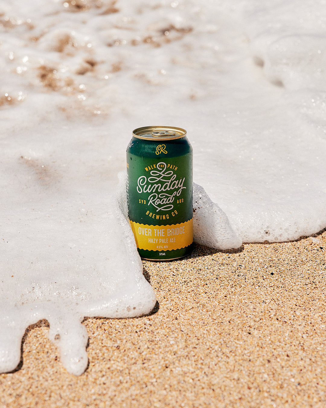 Can of Sunday Road Brewing beer on sand with ocean foam at wanda beach over the bridge hazy pale ale