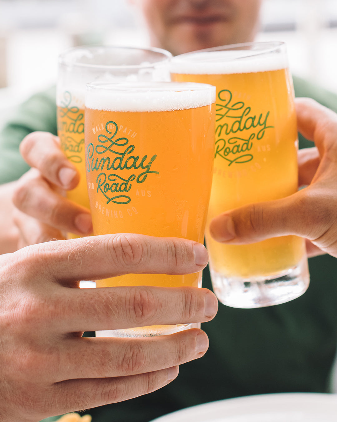 Three glasses of beer with 'Sunday Road' branding held by hands.