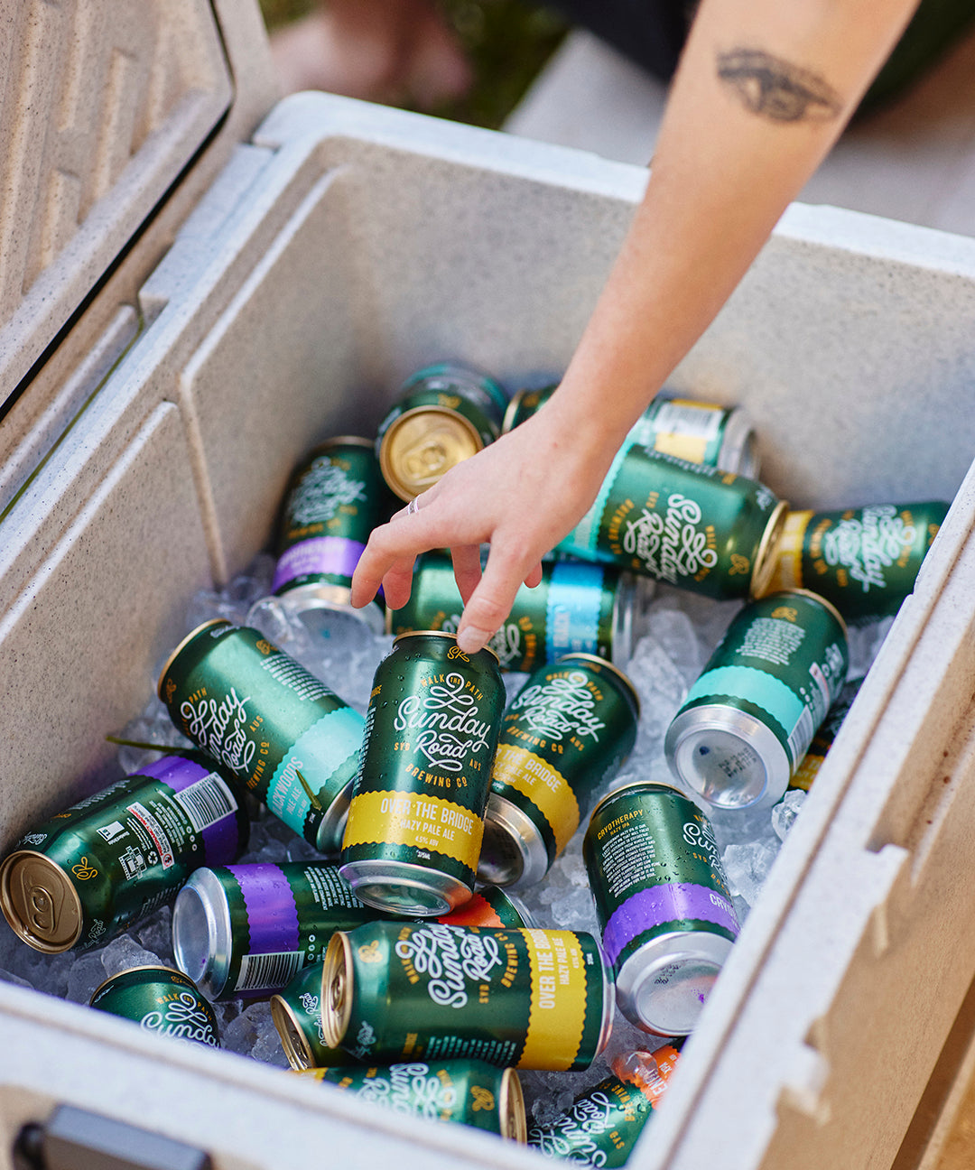Person reaching into a cooler filled with iced cans of sunday road beer from the sutherland shire