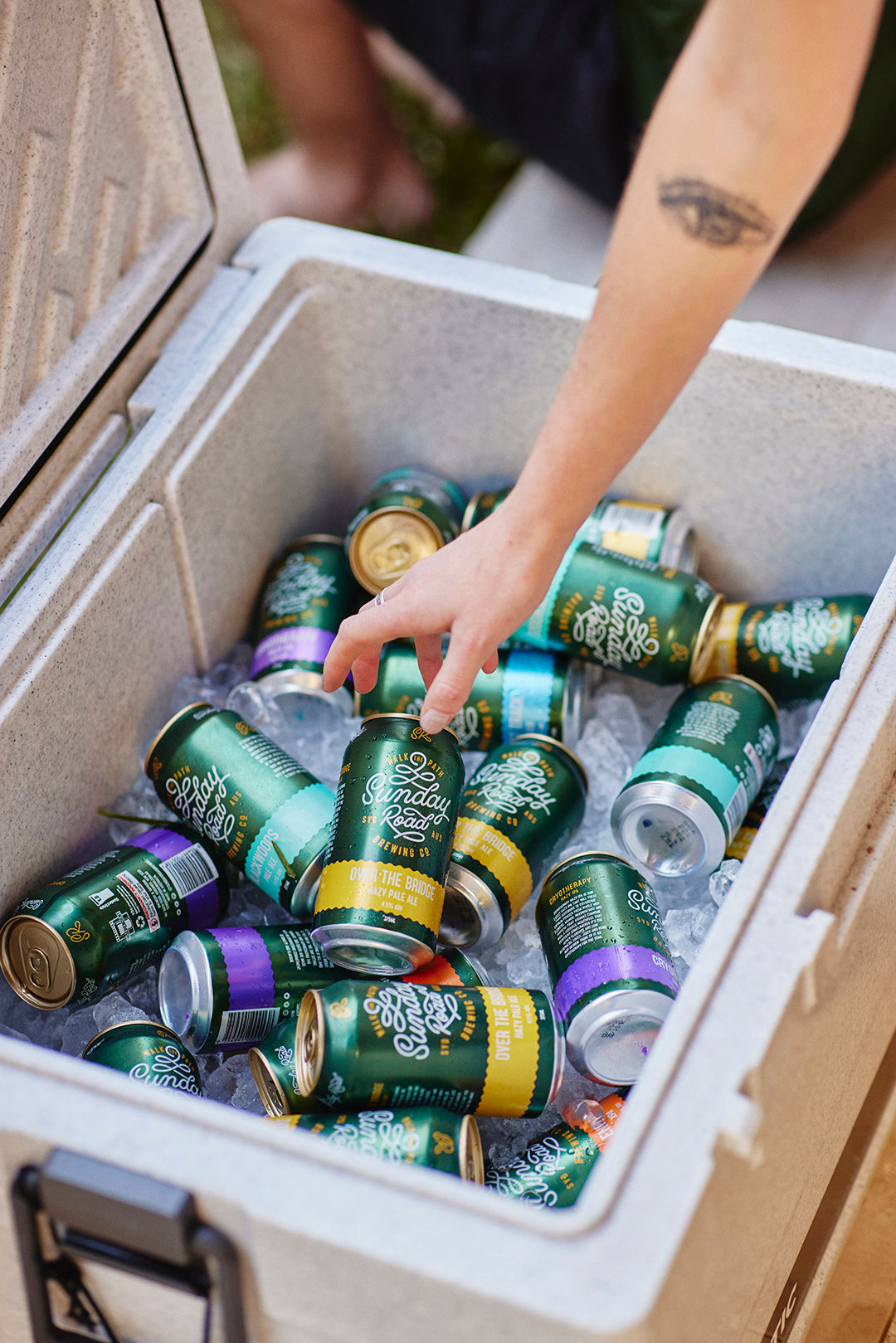 Person reaching into a cooler filled with iced cans of sunday road beer from the sutherland shire